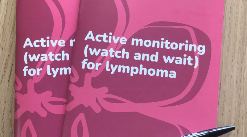 Two copies of the Active monitoring book, with a pen, on a wooden table. The book has a pink cover with periwinkle design and Lymphoma Action logo. Title of book is Active monitoring (watch and wait) for lymphoma.