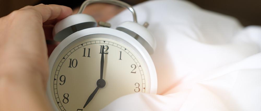 A person's hand holding an alarm clock, surrounded by bed sheets