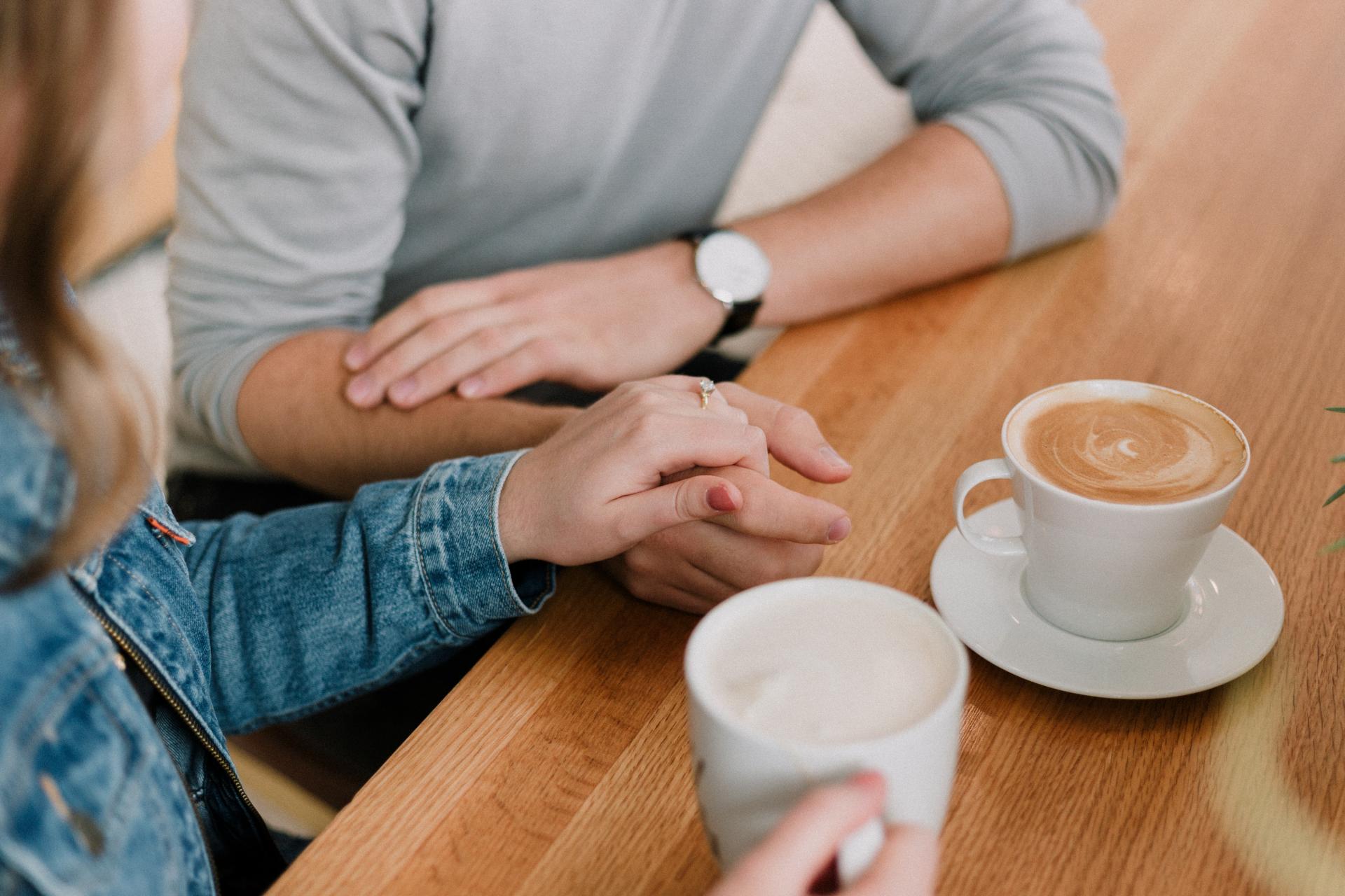 Two people sit at a table with cups of coffee, their hands touch