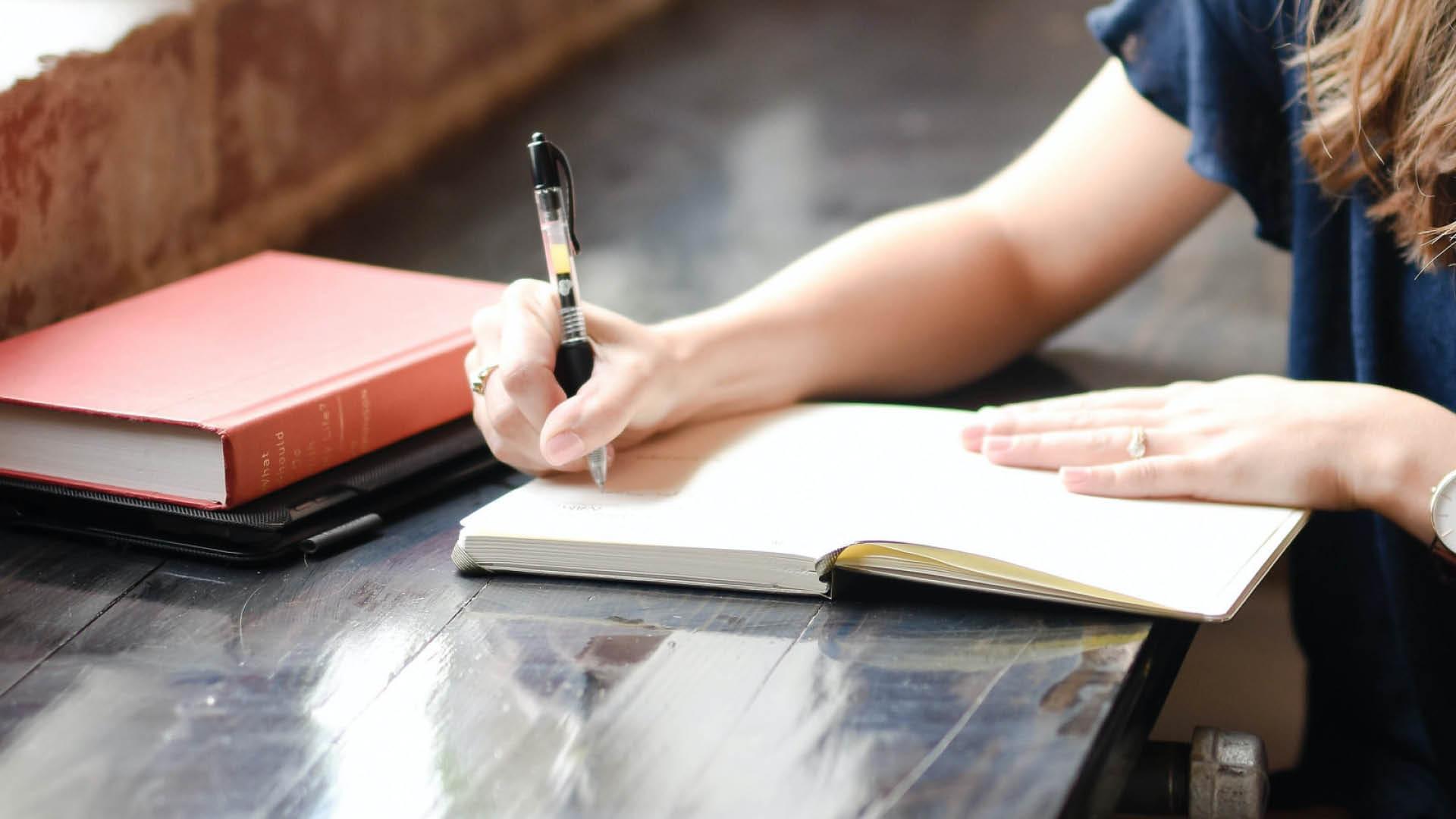 A person writes in a notebook, resting on a table. Other books are in the background.