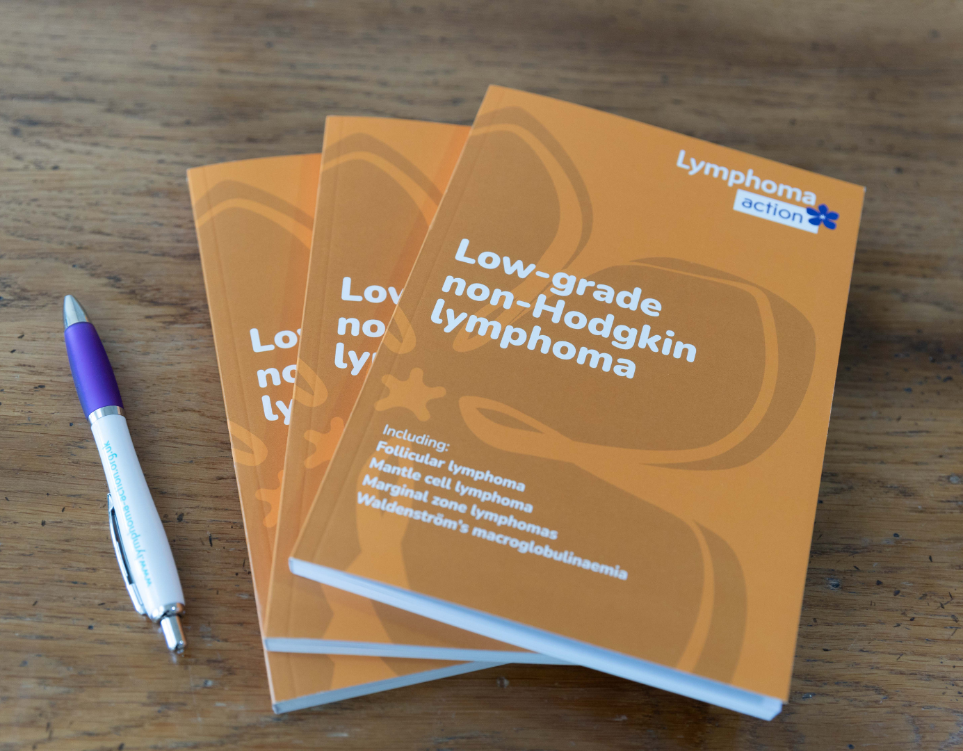 Image 3 copies of the book and a lymphoma action pen. Book has an orange cover with white writing. Title is Low-grade non-Hodgkin lymphoma. Lymphoma Action logo in top right-hand corner