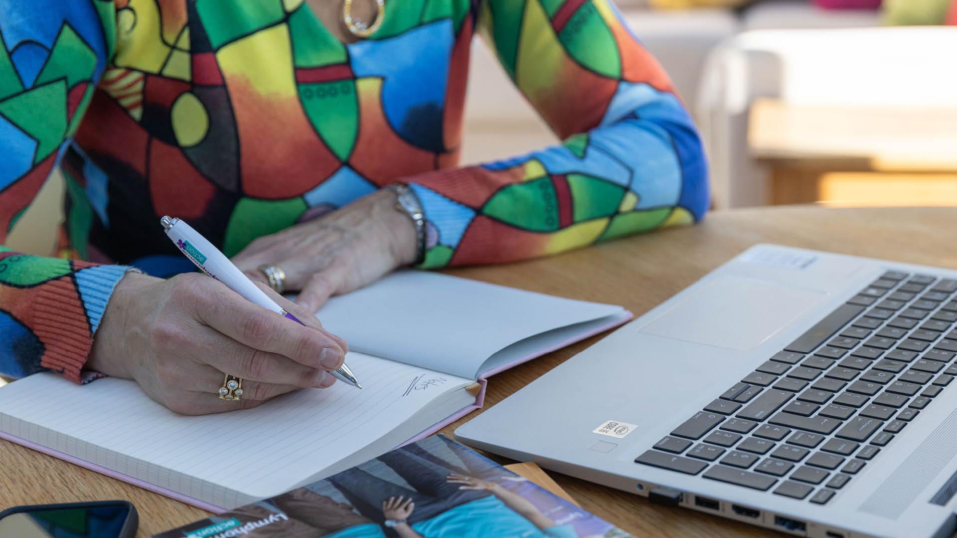 A person in a brightly coloured top is writing in a note book. A laptop is open beside her.