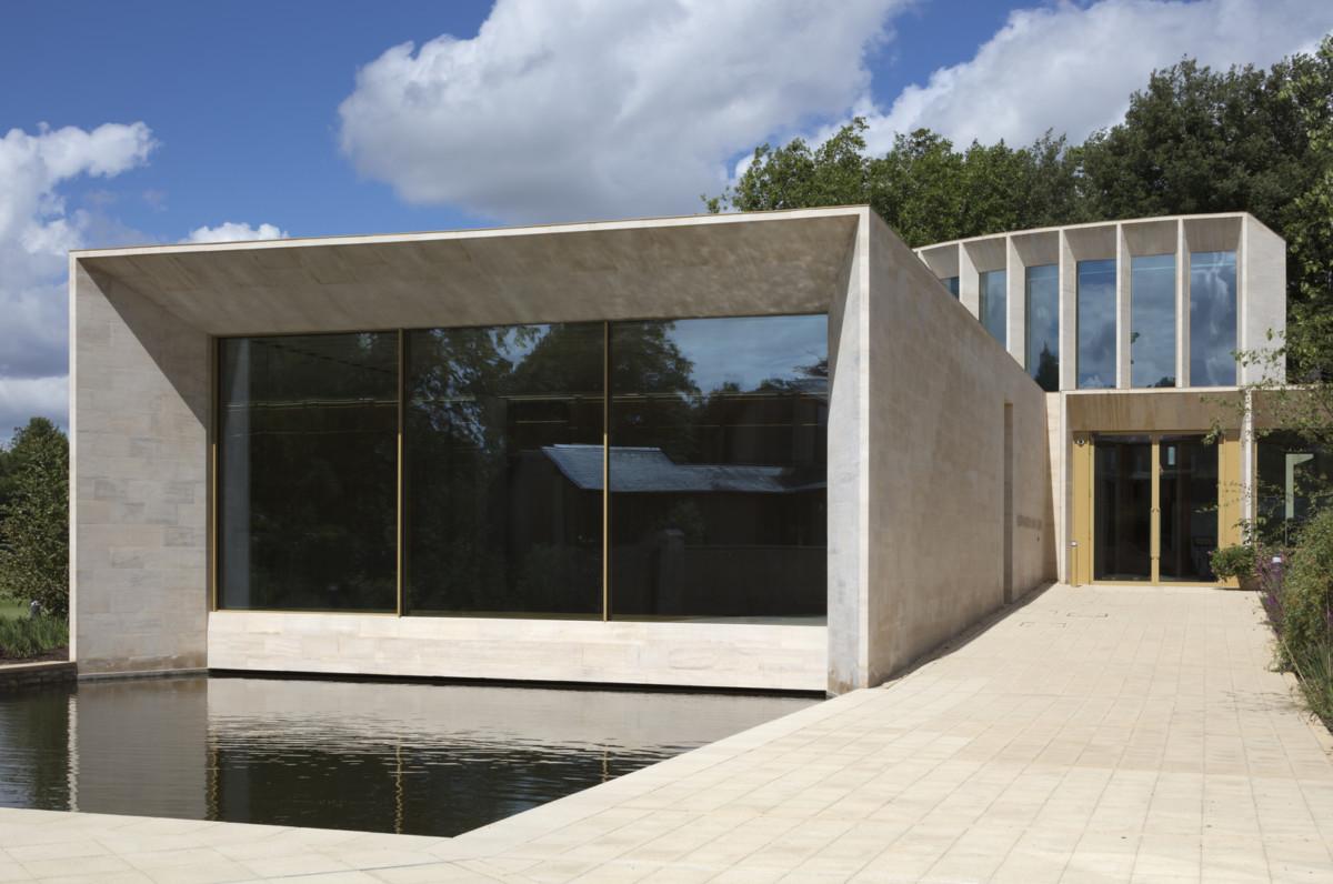 A square white buidling with large windows faces a water pool.