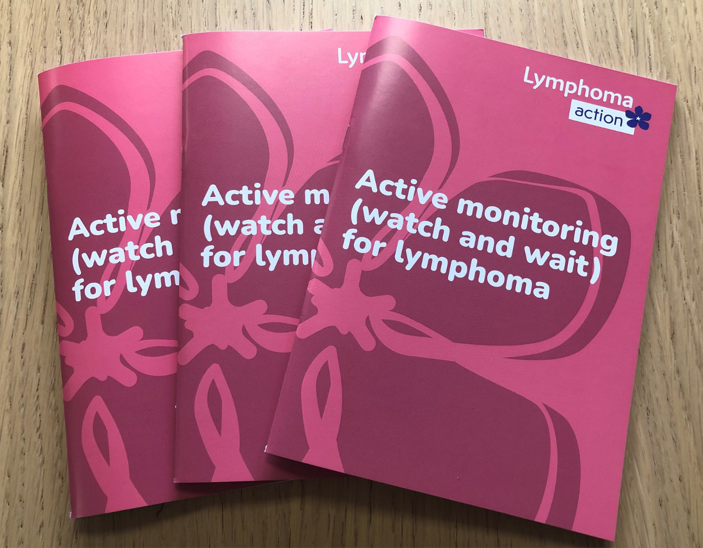 Three copies of the Active monitoring book on a wooden table. The book has a pink cover with periwinkle design and Lymphoma Action logo. Title of book is Active monitoring (watch and wait) for lymphoma.