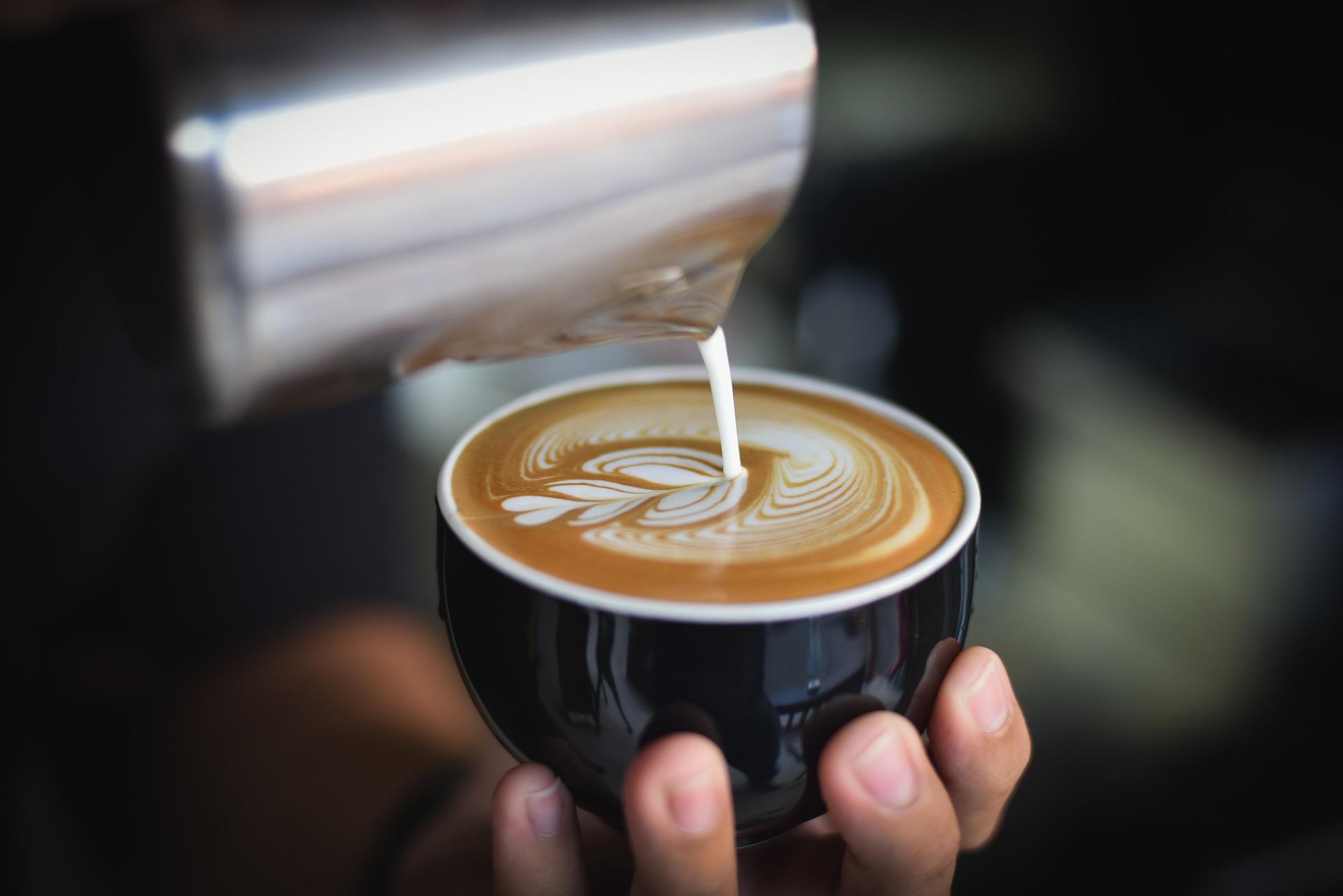 A hand holds a black cup as milk is poured into the coffee in a flower pattern