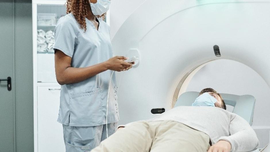 A nurse wearing a face mask talking to a person lying on a couch next to a donut-shaped scanner machine.