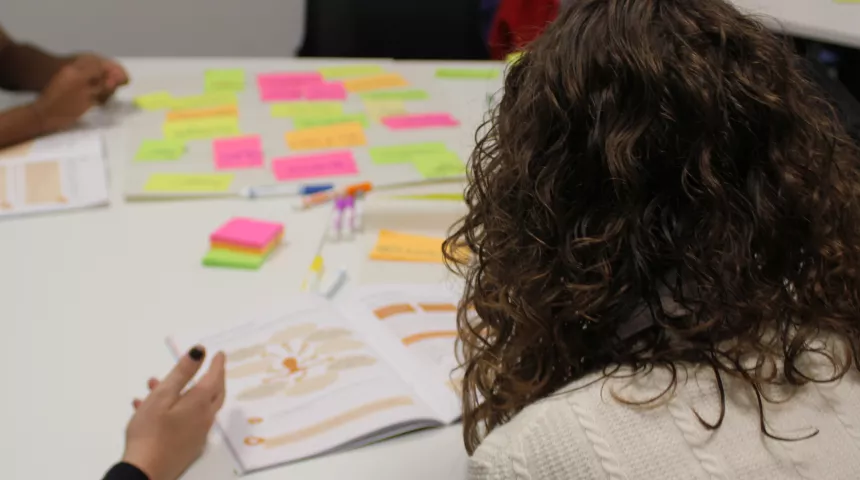 From behind, a person with long dark wavy hair leans over a white table with coloured sticky notes on it.
