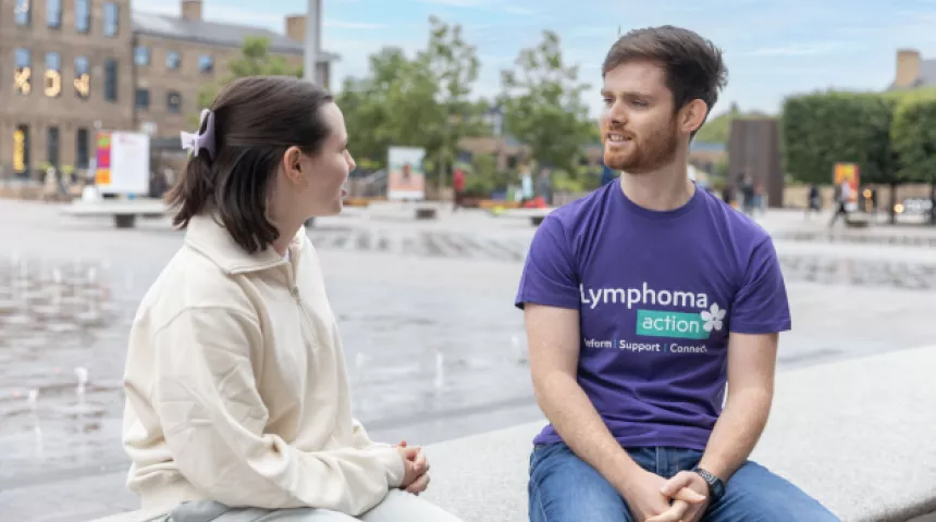 Two younger people sitting talking outside by a fountain with one of them wearing an LA t-shirt