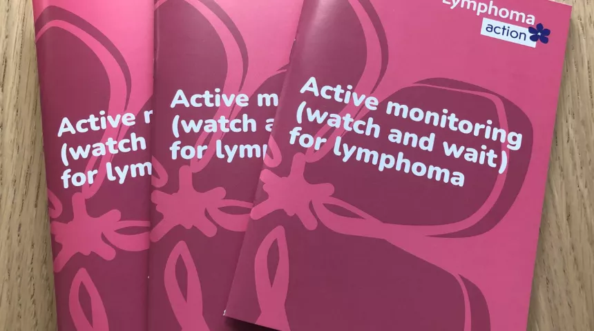 Three copies of the Active monitoring book on a wooden table. The book has a pink cover with periwinkle design and Lymphoma Action logo. Title of book is Active monitoring (watch and wait) for lymphoma.
