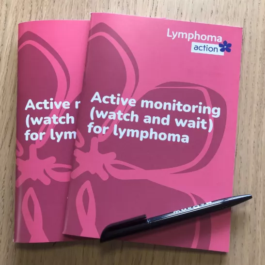 Two copies of the Active monitoring book, with a pen, on a wooden table. The book has a pink cover with periwinkle design and Lymphoma Action logo. Title of book is Active monitoring (watch and wait) for lymphoma.