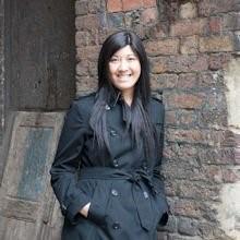 Young Asian woman in black coat leaning against a wall, smiling at the camera